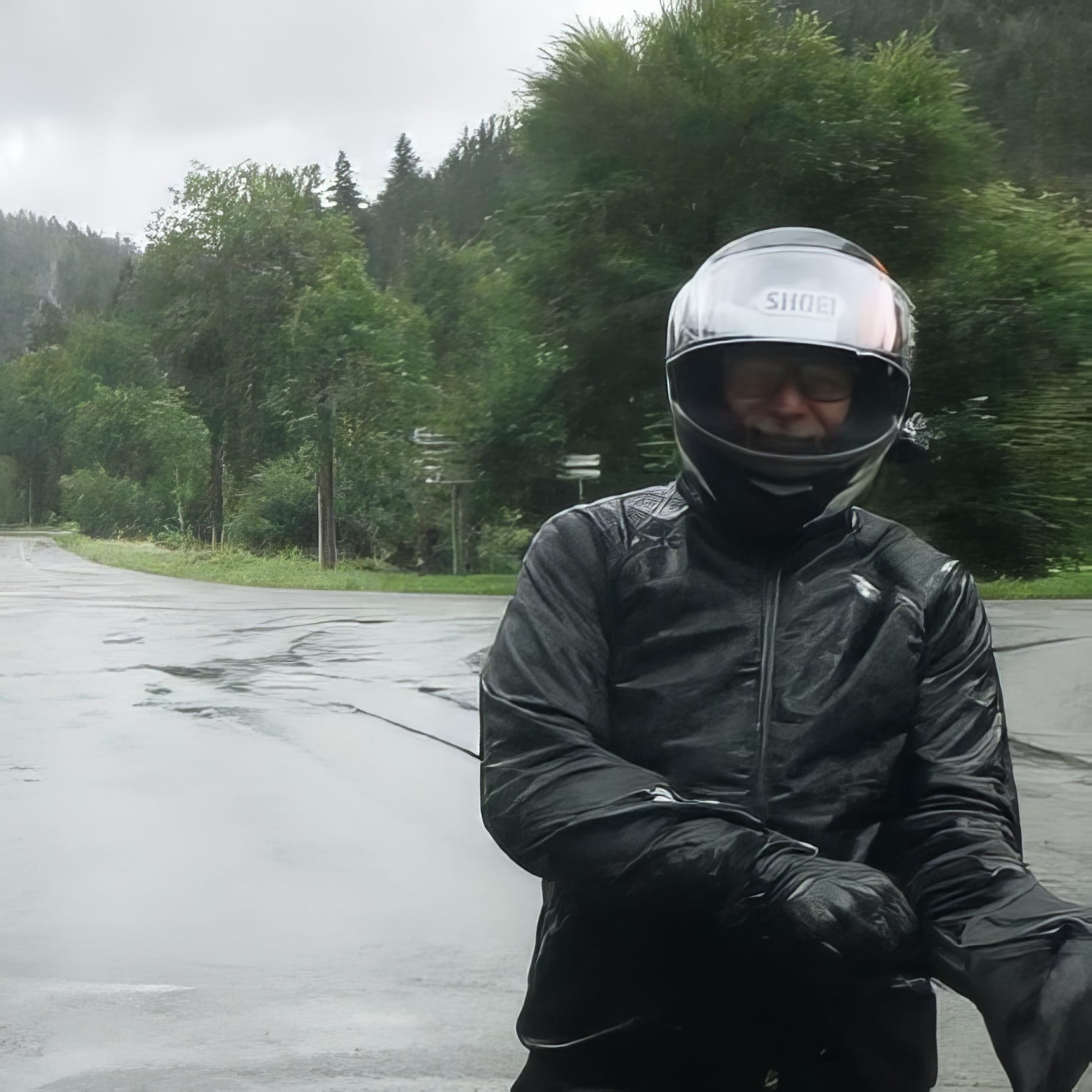 GPT  Das Foto zeigt einen Motorradfahrer in voller Schutzausrüstung. Er trägt einen schwarzen Helm, Jacke und Handschuhe. Der Hintergrund ist verschwommen, aber es scheint, als stünde der Fahrer auf einer nassen Straße, umgeben von grüner Vegetation, was auf regnerisches Wetter hindeutet. 