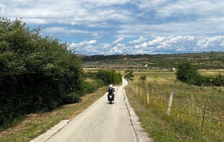 Ein Motorradfahrer fährt auf einer schmalen, ländlichen Strasse unter blauem Himmel mit weissen Wolken.