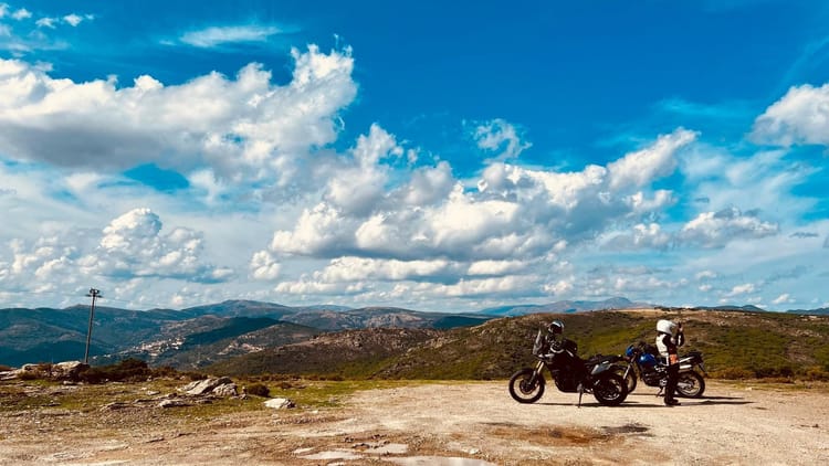 Zwei Motorräder und eine Person in Schutzkleidung in einer weiten, hügeligen Landschaft unter einem bewölkten, blauen Himmel.